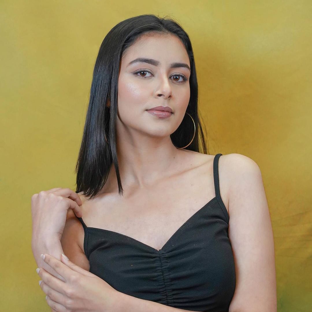 A woman named Rizul Singh, wearing a black top, striking a pose against a vibrant yellow background.